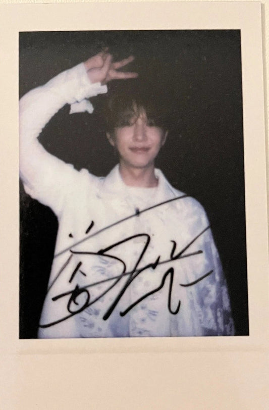 A signed polaroid-style photo print featuring a person in a white shirt, displayed on a light beige surface next to other printed photos.